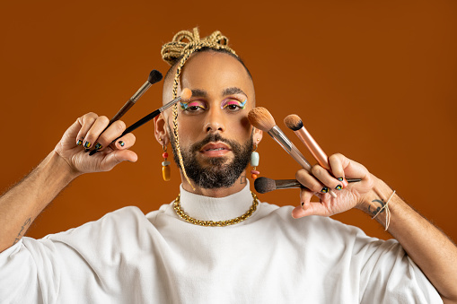 African american gay with bright make-up holding makeup brush in hand isolated on orange background. Handsome bearded make up artist. Fashion lgbtq concept.