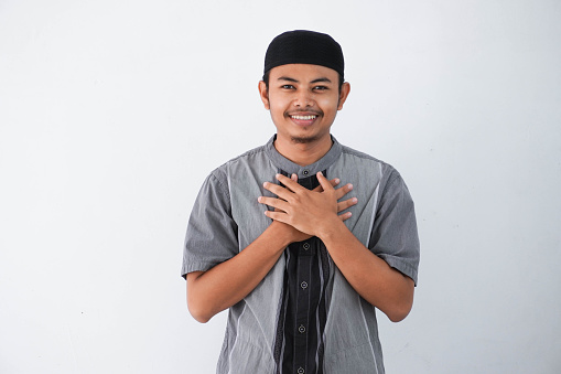 young Asian muslim man wearing grey muslim clothes with cap smiling with hands on chest with closed eyes and grateful gesture on face isolated on white background. health concept.