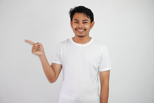 smiling asian man with fingers pointing to the side wearing white t shirt isolated on white background