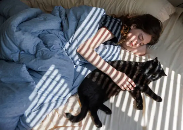 Photo of Striped shadows and lights in bedroom, bedtime relax: sleepy young woman hugging black cat in bed in sunbeams. Stripes of sunlight and shadow from the window cover bed and cat with owner. Top view