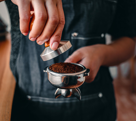 Close-up of hand barista or coffee maker holding portafilter and coffee tamper making an espresso coffee.