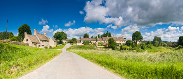Quiet country lane winding through lush green meadows to idyllic Cotswold hamlet of traditional limestone cottages under a panoramic summer sky. ProPhoto RGB profile for maximum color fidelity and gamut.