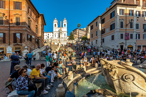 Rome, Italy - October 19, 2022: People sitting around Fontana della Barcaccia (Fountain of the longboat) on Piazza di Spagna as Spanish Steps on background in Rome - capital of Italy, famous and popular travel destination.