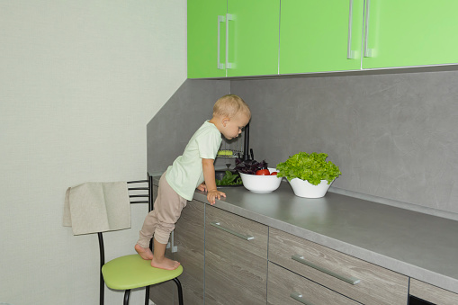 A little boy with a short haircut helps to cook in the kitchen. Washes fresh vegetables and herbs in a black sink in a gray kitchen...