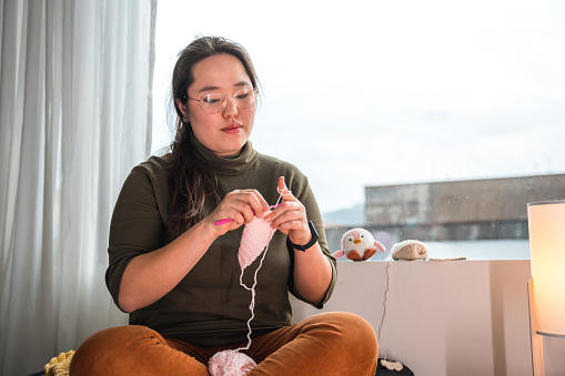 3/4 length image of a chubby Asian woman wearing glasses, sitting  comfortably on a sofa, her legs crossed, focusing on  knitting. Large window in the background.