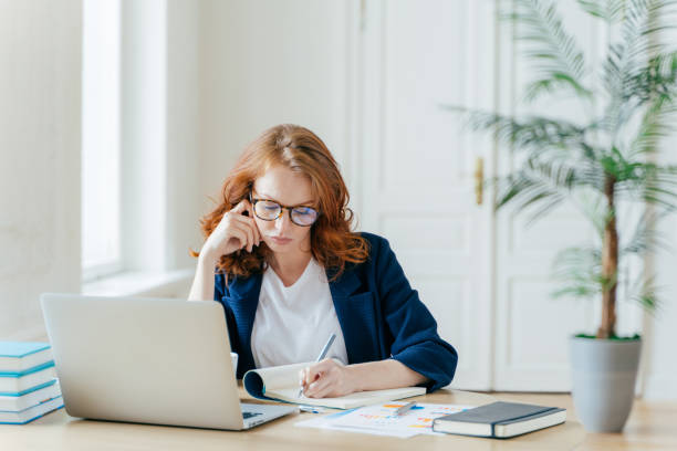istock Serious confident female entrepreneur in formal clothes writes down information while watches business webinar, notes main ideas, sits at work place in front of opened computer makes notes for startup 1489646782