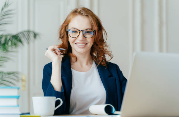 istock Glad redhead curly woman works freelance, uses laptop computer, writes down notes in notepad, drinks coffee, wears optical glasses, being in good mood, develops business company. Work concept 1489646811