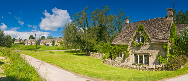 Traditional stone slate roofed cottage beside narrow country land leading though green meadows to picturesque Cotswold village under panoramic blue summer skies. ProPhoto RGB profile for maximum color fidelity and gamut.
