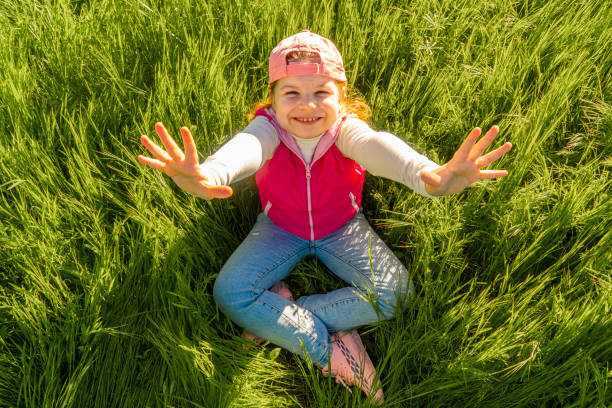 A cheerful girl sits in the thick green grass with her hands up in the air. Walk outdoors in the city park