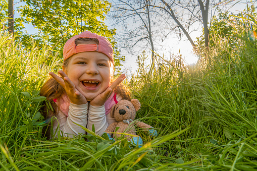 A cheerful girl lies in the thick green grass with a teddy bear against the backdrop of the sunset. Walk outdoors in the city park