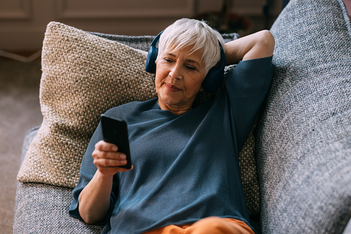 A from above view of a smiling senior Caucasian female enjoying listening to music on her smartphone while lying on the sofa in the living room.
