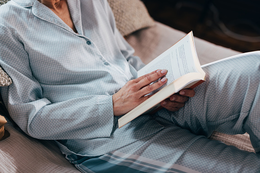 A close up view of an unrecognizable senior Caucasian female enjoying reading a book while sitting on bed in the bedroom.