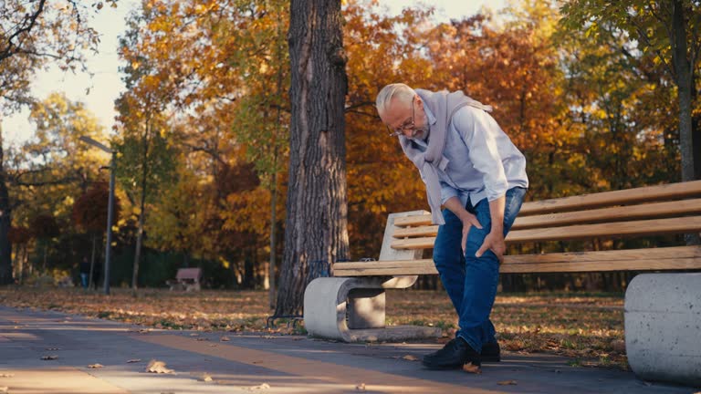 Senior man feeling knee pain when standing up from a bench in a park, injury