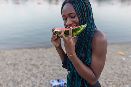 A photo of a young woman of African-American ethnicity near the water, enjoying a fresh watermelon on a sunny day.