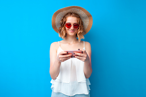 young girl in a sun hat and glasses use a smartphone on a blue isolated background, a woman in summer clothes looks at the phone