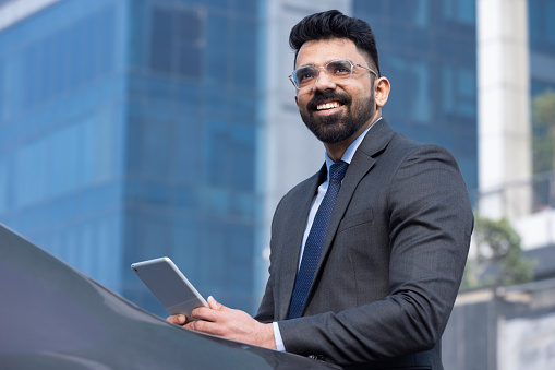 Stylish bearded businessman in formal business suit standing working with tablet in hands on background modern office building outside