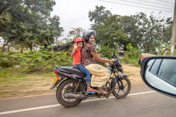 North Central Province, Sri Lanka - March 4th 2023:  Happy smiling son and mother driving on a motorcycle and smiling to the photographer in a passing car