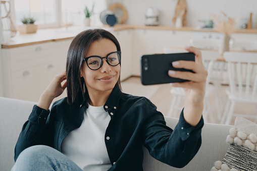 Hispanic girl is making selfie on smartphone. Woman is relaxing and using mobile phone camera. European lady enjoying time at home. Mobile phone, technologies and application using.