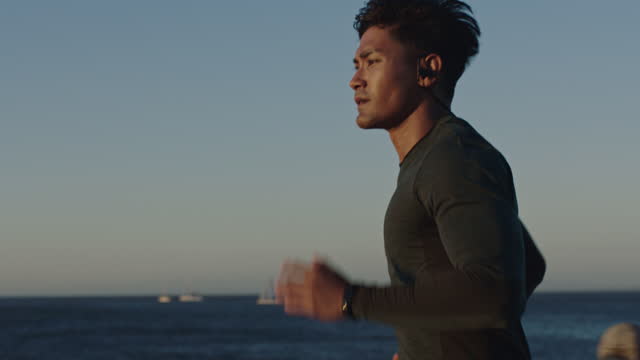 attractive young man running athlete endurance runner training jogging cardio exercise workout on calm ocean seaside run wearing earphones at sunset slow motion