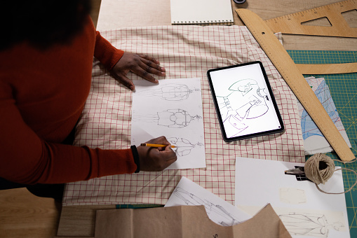 High angle view of unrecognizable African American female designer drawing sketches while looking at digital tablet in a clothing studio.