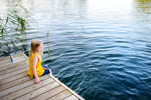 Cute little girl sitting on a wooden platform by the river or lake dipping her feet in the water on warm summer day