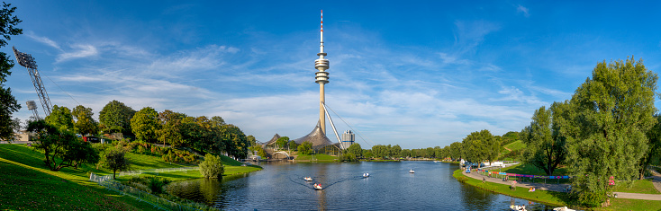 Panoramic photo of Olympic Park in Munich with Olympic Tower, lake, deciduous trees and blue sky in sunny summer weather