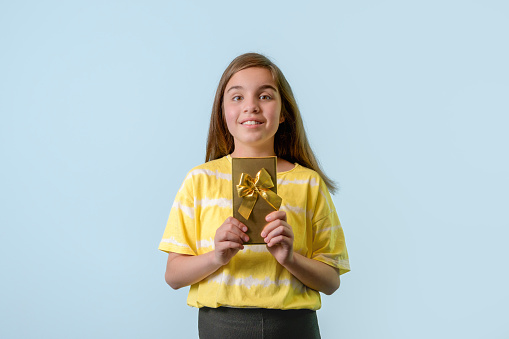 A joyful teenage girl holds a gift in a gilded package with a bow in her hands. Light blue background