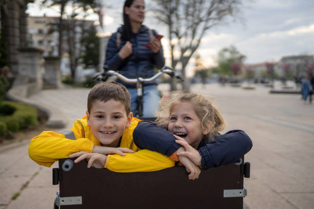 Little brother and sister having fun in cargo bicycle on the street city, their mother is sitting on a bicycle.