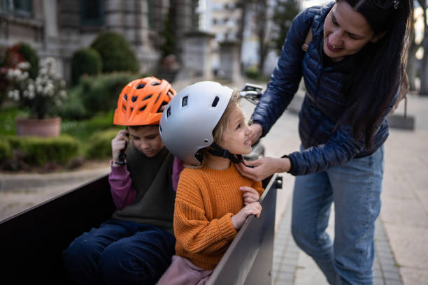 Mother putting cycling helmet on daughter head before ride in cargo bicycle in city.