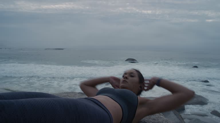 young hispanic woman runner doing sit ups exercise on beach at sunrise enjoying intense abs workout listenign to music for motivation athletic female enjoying fitness lifestyle