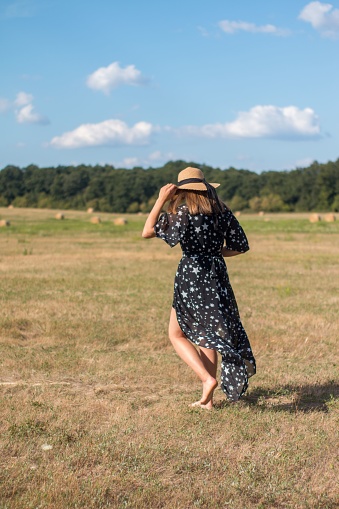 A young woman wearing a black dress and a straw hat walking in a meadow