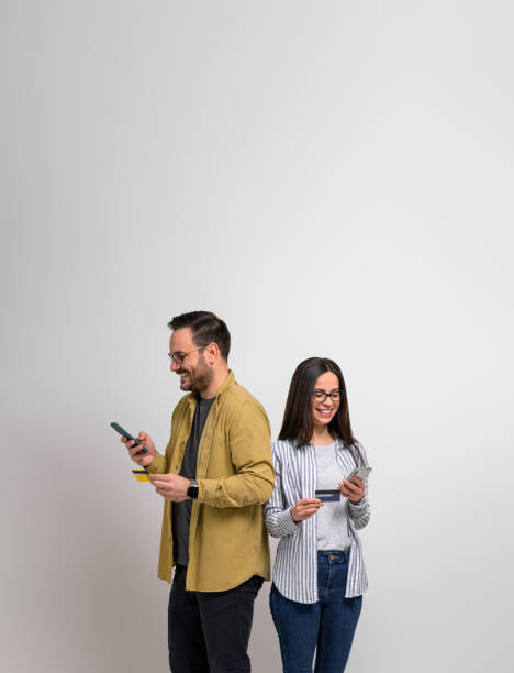 pareja joven sonriente comprando en línea con tarjeta de crédito y teléfonos inteligentes mientras está de pie contra el fondo. hombre y mujer felices comprando a través de redes sociales o aplicaciones de comercio electrónico - spending money fotos fotografías e imágenes de stock