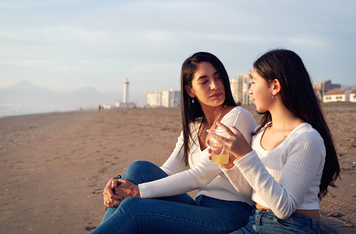 mid adult latina mother and daughter sitting on the beach talking and enjoying the sunset