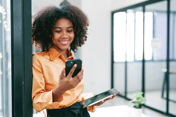 istock Young smiley black woman using mobile phone standing in office. 1489774441
