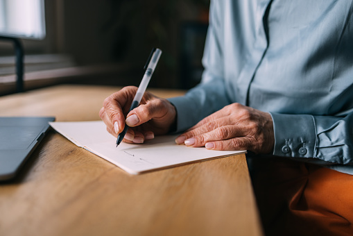 A close up view of an unrecognizable senior Caucasian female writing her diary while sitting at the desk in  front of her laptop.