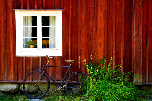 A Vintage bike is leaning against a red, wooden House Wall with a Window in the left
