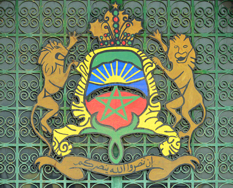 National coat of arms of the Kingdom of Morocco - introduced in 1957 when Mohammed V was proclaimed king of Morocco. Features a green pentagram on red and a rising sun, above it the royal crown. Two lions act as shield holders. On the band below the Arabic language inscription: ' If you help God, he will also help you' (Koran, verse 7, Surah 47) - building exterior.