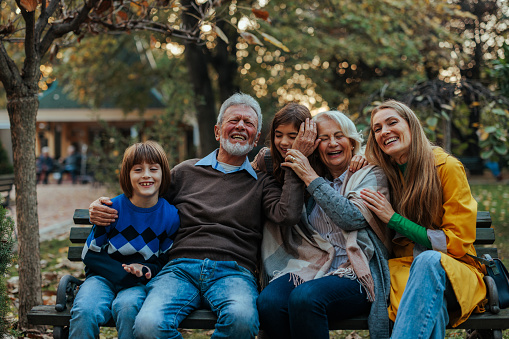 A multigenerational family is in the park sitting on the bench having a big hug as they enjoy their leisure time outdoors.