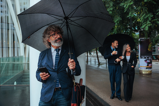 A businessman is standing on the street on a rainy day with an umbrella and using his cellphone.