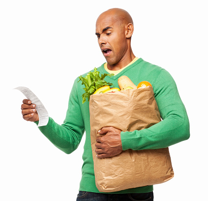 Young African American man in casual wear staring at bill in disbelief while holding grocery bag. Horizontal shot. Isolated on white.