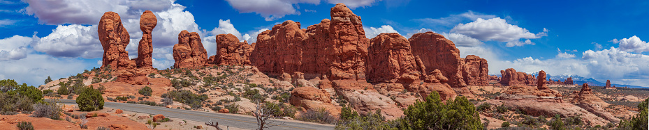 A panoramic view of the road running along the incredible shape of the rock formations in Arches National Park, and beautiful clouds in the sky highlight the beauty of these places.