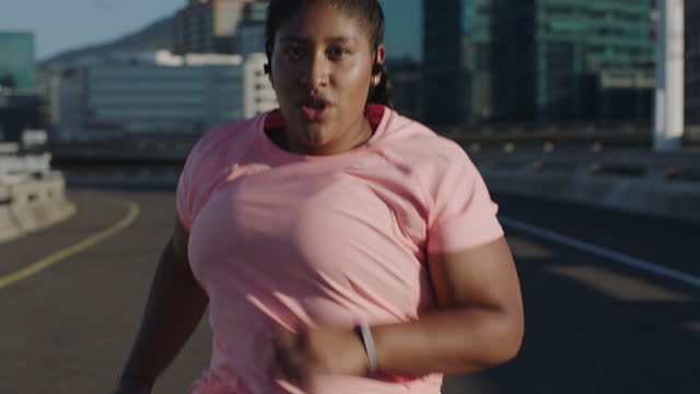 fat african american woman running exercising intense weight loss workout jogging in urban city background at sunrise wearing earphones
