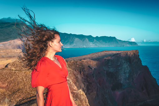 Description: Woman in red dress enjoys the raw, windy atmosphere above the foothills of a volcanic island in the Old Atlantic Ocean. São Lourenço, Madeira Island, Portugal, Europe.