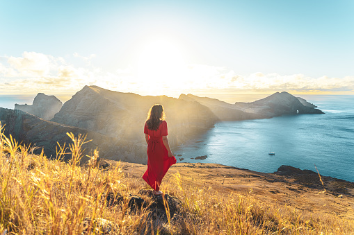 Description: Woman in red dress stands on rock in yellow field enjoying the scenery above the foothills of a volcanic island in the Atlantic Ocean. São Lourenço, Madeira Island, Portugal, Europe.