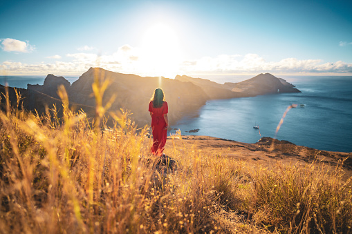 Description: Woman in red dress sunbathing with beautiful view on the foothills of a volcanic island in the Atlantic Ocean. São Lourenço, Madeira Island, Portugal, Europe.