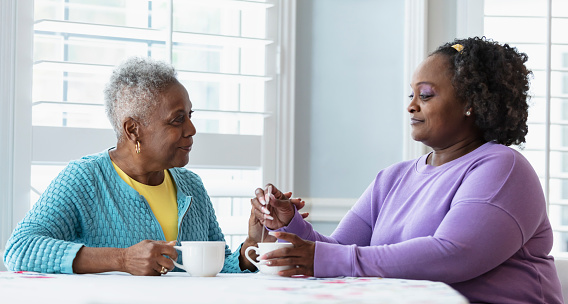 A senior African-American woman sitting at home at a table, having tea with her adult daughter. They look relaxed, and the mother's hand is resting affectionately on her daughter's.