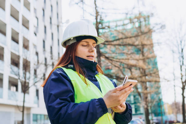 istock Architect woman in white hardhat and safety vest with digital tablet using smartphone outdoors. Female engineer. Logistics and construction worker near modern building. Engineering application 1489847613