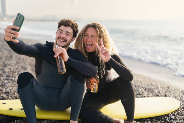 Couple of surfers taking selfie with mobile phone, while taking break from surfing stock photo