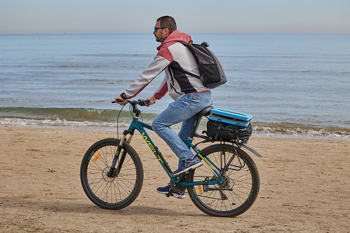 A man with a backpack on the beach rides a bicycle on Malvarosа beach in Valencia.