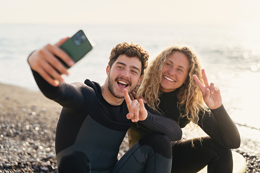 Young Caucasian woman and her male surf instructor, taking selfie with mobile phone, while sitting on the surfboard